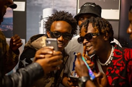 A group of young men are gathered closely together, all wearing stylish attire and sunglasses. They appear to be looking at something on a smartphone with expressions of excitement and amusement. The environment seems casual and social.