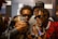 A group of young men are gathered closely together, all wearing stylish attire and sunglasses. They appear to be looking at something on a smartphone with expressions of excitement and amusement. The environment seems casual and social.