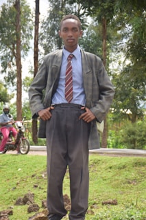 A young person stands outdoors, wearing a suit with a striped tie. The background includes tall trees and a person riding a motorcycle. The setting appears to be a lush, green area with grass and a road.