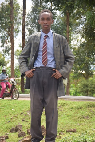 A young person stands outdoors, wearing a suit with a striped tie. The background includes tall trees and a person riding a motorcycle. The setting appears to be a lush, green area with grass and a road.