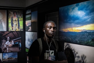 A man wearing headphones stands in a dimly lit room with art displayed on the walls. The room features several framed photographs, including a vivid image of a dramatic sunset over rolling hills and portraits of various individuals.