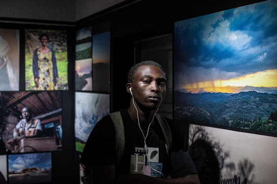A man wearing headphones stands in a dimly lit room with art displayed on the walls. The room features several framed photographs, including a vivid image of a dramatic sunset over rolling hills and portraits of various individuals.