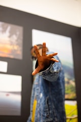 A person wearing a blue denim jacket is extending their hand towards the camera, with three fingers spread out. Several framed photographs or pictures are mounted on the wall behind them, depicting various scenes including a landscape and a sunset.