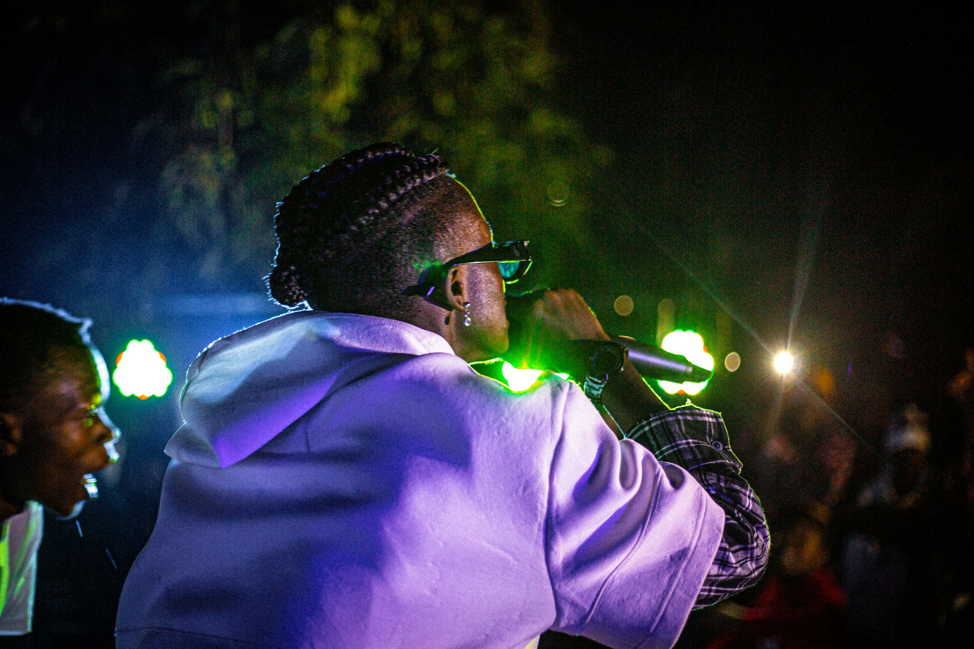 A person wearing glasses and a white hoodie is holding a microphone on a stage. They have braided hair and are illuminated by stage lights. Another person is partially visible on the left, looking towards the performer. The background is dark with colorful lights creating a vibrant atmosphere.
