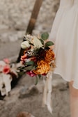 A close-up of a floral bouquet held by a person wearing a white garment. The bouquet features a variety of flowers in warm colors, including oranges, reds, and yellows, complemented by green leaves and some dried elements. The background is blurred, with faint hints of another floral arrangement and an outdoor setting.