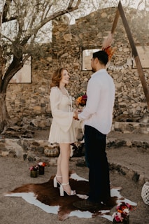 A couple is standing facing each other in an outdoor setting with rustic stone walls in the background. The woman is wearing a short white dress and high heels, holding a bouquet of flowers. The man is dressed in a white shirt and dark pants. They are standing on an animal hide laid on the ground, surrounded by small floral arrangements and decor. Sunlight filters through the tree branches giving the scene a warm, intimate atmosphere.
