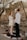 A couple is standing facing each other in an outdoor setting with rustic stone walls in the background. The woman is wearing a short white dress and high heels, holding a bouquet of flowers. The man is dressed in a white shirt and dark pants. They are standing on an animal hide laid on the ground, surrounded by small floral arrangements and decor. Sunlight filters through the tree branches giving the scene a warm, intimate atmosphere.