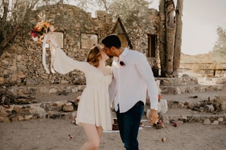 A couple is embracing and kissing outdoors in front of a rustic stone building. The woman is wearing a white dress and holding a bouquet of brightly colored flowers, while the man is in a white shirt. The setting is desert-like, with cacti and dry earth visible.