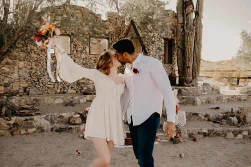 A couple is embracing and kissing outdoors in front of a rustic stone building. The woman is wearing a white dress and holding a bouquet of brightly colored flowers, while the man is in a white shirt. The setting is desert-like, with cacti and dry earth visible.