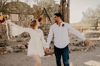 A couple is holding hands and smiling at each other while walking outdoors, surrounded by stone ruins and desert vegetation. The woman is wearing a white dress and holding a bouquet of flowers. The man is wearing a white shirt with a red floral decoration on his chest and dark jeans. The scenery includes an old stone wall, a wooden structure in the background, and a dry, sandy ground.