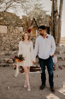 A couple standing hand in hand in a rustic outdoor setting with stone walls and desert plants. The woman is wearing a light cream dress and holding a bouquet of flowers, while the man is dressed in a white shirt and jeans. The atmosphere is casual yet intimate, with floral arrangements and candles placed around.