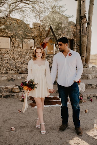 A couple standing hand in hand in a rustic outdoor setting with stone walls and desert plants. The woman is wearing a light cream dress and holding a bouquet of flowers, while the man is dressed in a white shirt and jeans. The atmosphere is casual yet intimate, with floral arrangements and candles placed around.
