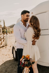 A couple embraces affectionately in a natural setting with a white trailer behind them. The woman holds a colorful bouquet of flowers, and both are dressed in casual yet elegant attire. The sun casts a warm glow over the scene, and the background includes desert vegetation and distant hills.