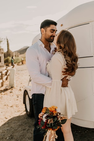 A couple embraces affectionately in a natural setting with a white trailer behind them. The woman holds a colorful bouquet of flowers, and both are dressed in casual yet elegant attire. The sun casts a warm glow over the scene, and the background includes desert vegetation and distant hills.