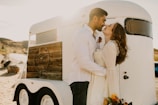 A couple stands in an affectionate embrace beside a vintage-style trailer. Sunlight softly illuminates the scene, casting a warm glow. The woman, holding a colorful bouquet, gazes lovingly at the man, who wears a white shirt with a floral boutonniere.