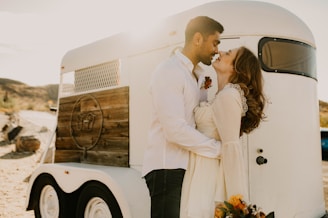 A couple stands in an affectionate embrace beside a vintage-style trailer. Sunlight softly illuminates the scene, casting a warm glow. The woman, holding a colorful bouquet, gazes lovingly at the man, who wears a white shirt with a floral boutonniere.