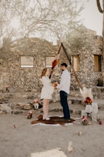 A couple stands facing each other in a rustic outdoor setting. They are holding flowers, surrounded by decorative items such as vases, pampas grass, and small floral arrangements. The background features a stone wall and a wooden triangle arch adorned with foliage. The scene has a natural, earthy ambiance.