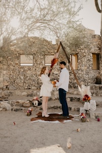 A couple stands facing each other in a rustic outdoor setting. They are holding flowers, surrounded by decorative items such as vases, pampas grass, and small floral arrangements. The background features a stone wall and a wooden triangle arch adorned with foliage. The scene has a natural, earthy ambiance.
