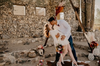 A couple shares a passionate kiss in an outdoor setting decorated for a rustic wedding. The man is wearing a white shirt and jeans, while the woman is in a short, white dress and heels. They are positioned under a triangular arch adorned with dried flowers. The background features a stone wall and natural elements such as cacti and various floral arrangements.