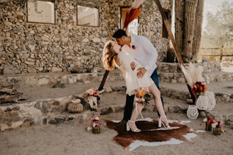 A couple shares a romantic kiss in an outdoor setting with a rustic stone building in the background. The woman is wearing a short white dress and heels, while the man is dressed in a white shirt and jeans. They stand on an animal hide rug, surrounded by decorative elements like pampas grass, flowers, and lanterns.