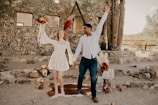 A joyful couple is holding hands and raising their free arms in celebration. They are surrounded by a rustic setting with stone walls and cacti in the background. The woman wears a white dress and holds a colorful bouquet. Various decorative elements, including flowers and lanterns, are arranged around them.