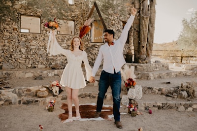 A joyful couple is holding hands and raising their free arms in celebration. They are surrounded by a rustic setting with stone walls and cacti in the background. The woman wears a white dress and holds a colorful bouquet. Various decorative elements, including flowers and lanterns, are arranged around them.