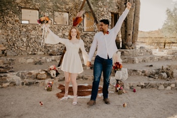 A couple is joyfully posing in front of a rustic stone building. The woman is wearing a short white dress and holding a bouquet of flowers, while the man is dressed in a white shirt and jeans. Various decorative elements like flowers and lanterns are arranged around them, and a large cactus stands nearby.
