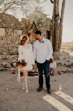 A couple stands hand in hand in a rustic outdoor setting. The woman is wearing a white dress and holds a bouquet of colorful flowers. The man is dressed in a white shirt with a flower pinned to it. They are surrounded by a stone structure, desert plants, and various decorations on the ground. The natural sunlight casts soft shadows, creating a warm atmosphere.