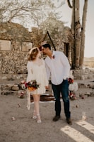 A couple stands in a rustic outdoor setting, near a stone structure with a large cactus nearby. The woman is wearing a light-colored dress and holding a bouquet of flowers, while the man is in a white shirt and dark jeans. Floral arrangements and decorative elements are placed around them, creating a romantic atmosphere.