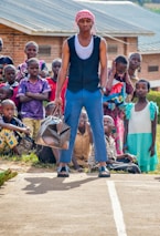 A group of children are gathered outdoors with a focused young man standing in the center, holding a patterned travel bag. He wears a red and white headscarf, a sleeveless shirt, blue pants, and sandals. The children behind him display varied expressions, and some are seated on the ground. A brick building with a metal roof is visible in the background.
