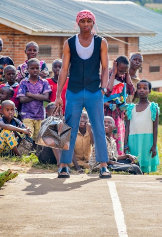 A group of children are gathered outdoors with a focused young man standing in the center, holding a patterned travel bag. He wears a red and white headscarf, a sleeveless shirt, blue pants, and sandals. The children behind him display varied expressions, and some are seated on the ground. A brick building with a metal roof is visible in the background.