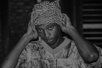 A person with a serious expression adjusts a patterned headscarf with both hands. They are wearing a camouflage-patterned shirt, and the background is blurred, making the individual the focal point.