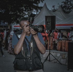 A person is taking a photograph with a professional Nikon camera while standing outdoors among a crowd of people. The person is wearing a dark vest with pockets, and tents with red and white stripes are visible in the background. There is also a large black speaker and several attendees visible, some wearing pink or light-colored clothing.