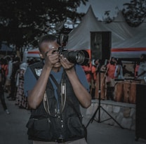 A person is taking a photograph with a professional Nikon camera while standing outdoors among a crowd of people. The person is wearing a dark vest with pockets, and tents with red and white stripes are visible in the background. There is also a large black speaker and several attendees visible, some wearing pink or light-colored clothing.