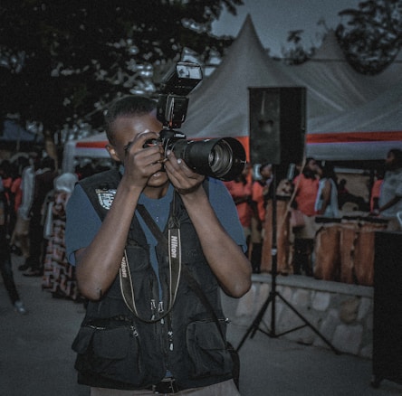 A person is taking a photograph with a professional Nikon camera while standing outdoors among a crowd of people. The person is wearing a dark vest with pockets, and tents with red and white stripes are visible in the background. There is also a large black speaker and several attendees visible, some wearing pink or light-colored clothing.