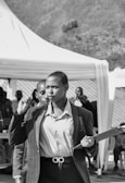 A person in business attire is holding a pen and a clipboard, walking purposefully with a focused expression. In the background, there is a gathering of people and a tent set up outdoors.