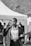 A person in business attire is holding a pen and a clipboard, walking purposefully with a focused expression. In the background, there is a gathering of people and a tent set up outdoors.