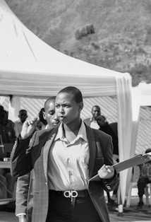 A person in business attire is holding a pen and a clipboard, walking purposefully with a focused expression. In the background, there is a gathering of people and a tent set up outdoors.