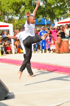 A young performer is captured mid-dance, wearing a white shirt and black pants, displaying a high kick. An engaged audience is visible in the background, including people in vibrant clothing and cultural attire. The scene takes place outdoors, with sunny weather and trees providing a natural backdrop.
