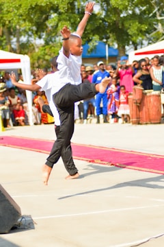 A young performer is captured mid-dance, wearing a white shirt and black pants, displaying a high kick. An engaged audience is visible in the background, including people in vibrant clothing and cultural attire. The scene takes place outdoors, with sunny weather and trees providing a natural backdrop.