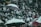 A diverse group of people are gathered, sitting closely together in a stadium-like setting. Many are smiling and clapping, creating an atmosphere of enthusiasm and camaraderie. Umbrellas are opened among the crowd, suggesting outdoor seating and possibly providing shade or protection from rain.
