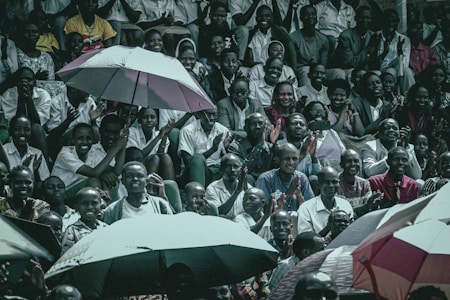 A diverse group of people are gathered, sitting closely together in a stadium-like setting. Many are smiling and clapping, creating an atmosphere of enthusiasm and camaraderie. Umbrellas are opened among the crowd, suggesting outdoor seating and possibly providing shade or protection from rain.
