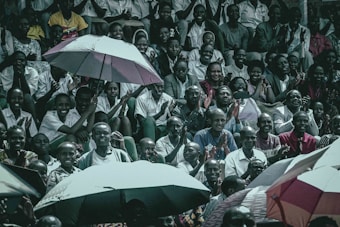 A diverse group of people are gathered, sitting closely together in a stadium-like setting. Many are smiling and clapping, creating an atmosphere of enthusiasm and camaraderie. Umbrellas are opened among the crowd, suggesting outdoor seating and possibly providing shade or protection from rain.