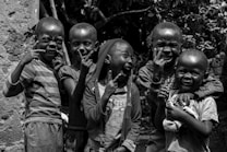 A group of five children express joy and excitement while standing outdoors. Each child is posing playfully, with wide smiles and gestures. The setting appears natural, with foliage visible in the background.