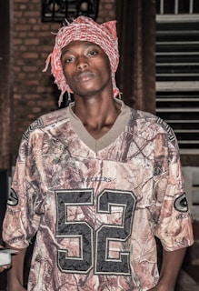A person is wearing a camouflage-patterned Packers jersey with the number 52 prominently displayed. They have a red and white patterned headscarf wrapped around their head. The background consists of a brick wall and a set of blinds or shutters on the right side.