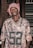 A person is wearing a camouflage-patterned Packers jersey with the number 52 prominently displayed. They have a red and white patterned headscarf wrapped around their head. The background consists of a brick wall and a set of blinds or shutters on the right side.