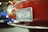 A close-up of a classic red car with a Michigan license plate that reads 'AMPHICAR'. The car's headlight and chrome bumper are visible, along with a blurred blue vehicle in the background.
