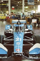A blue and black race car is prominently displayed indoors, surrounded by vintage cars and motorcycles. The car has the number 21 on it and logos indicating sponsorship from Honda and Chevrolet. Two people are visible in the background, observing the exhibits.
