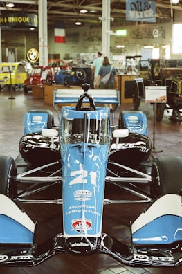 A blue and black race car is prominently displayed indoors, surrounded by vintage cars and motorcycles. The car has the number 21 on it and logos indicating sponsorship from Honda and Chevrolet. Two people are visible in the background, observing the exhibits.