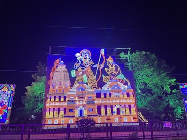 An illuminated decoration depicts two epic figures, likely from mythology, holding a bow and arrow. The scene includes a large temple-like structure adorned with vibrant, colorful lights. Trees in the background are also lit up, adding to the festive atmosphere.